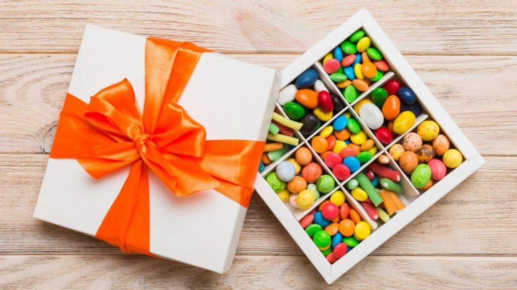 A white gift box with an orange ribbon is open to reveal assorted colorful candies organized in a grid of nine compartments, set on a light wooden surface—perfectly capturing the charm of a Route 66 candy box.