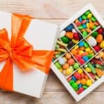 A white gift box with an orange ribbon is open to reveal assorted colorful candies organized in a grid of nine compartments, set on a light wooden surface—perfectly capturing the charm of a Route 66 candy box.