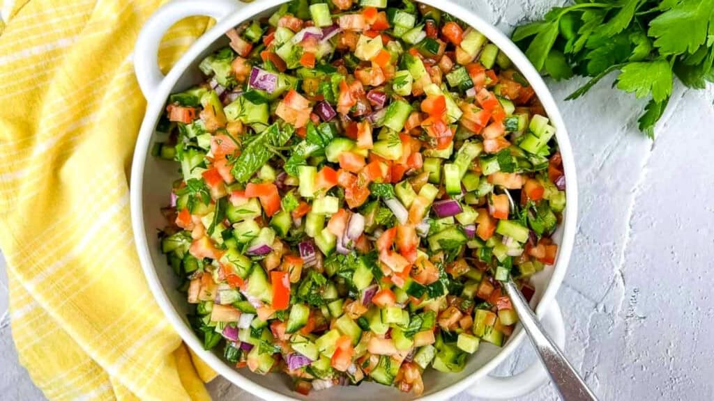 A white bowl filled with chopped salad containing cucumber, tomato, red onion, and herbs, with a spoon inside. A yellow towel and green herbs are beside the bowl.