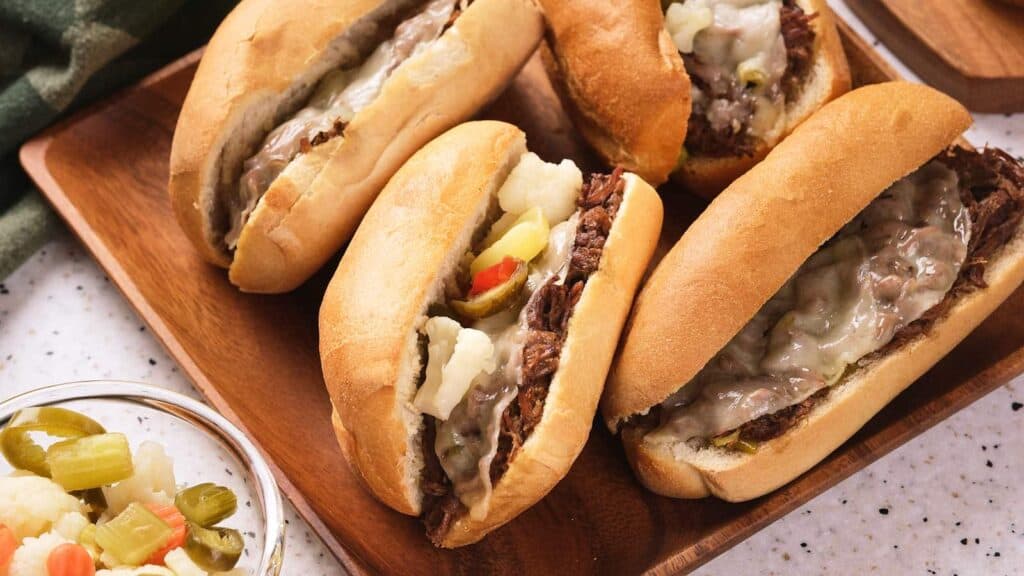 Four beef sandwiches with melted cheese and pickled vegetables on a wooden tray, with a small bowl of additional pickled vegetables on the side.