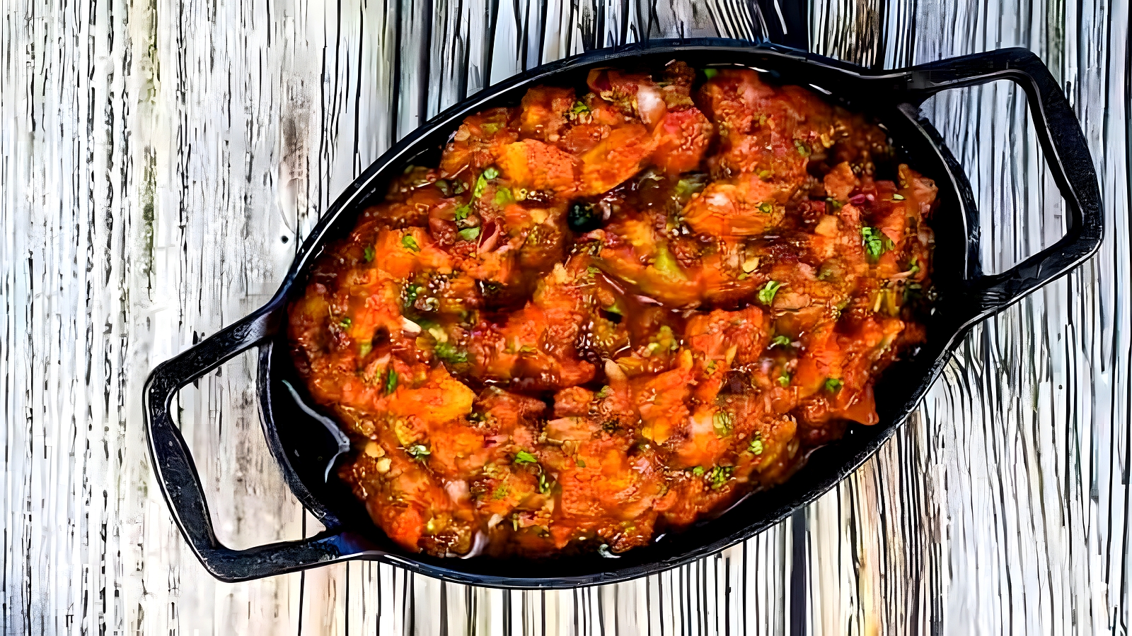 Black oval dish filled with a chunky red-brown stew or curry, garnished with green herbs, placed on a wooden surface.