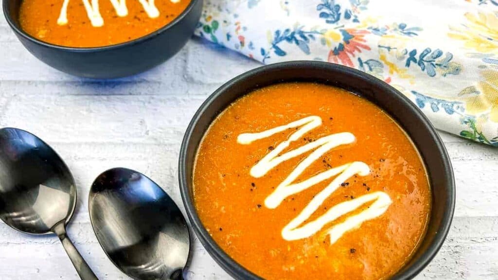 Two bowls of tomato soup topped with a swirl of cream, next to two metal spoons on a white surface with a floral napkin.