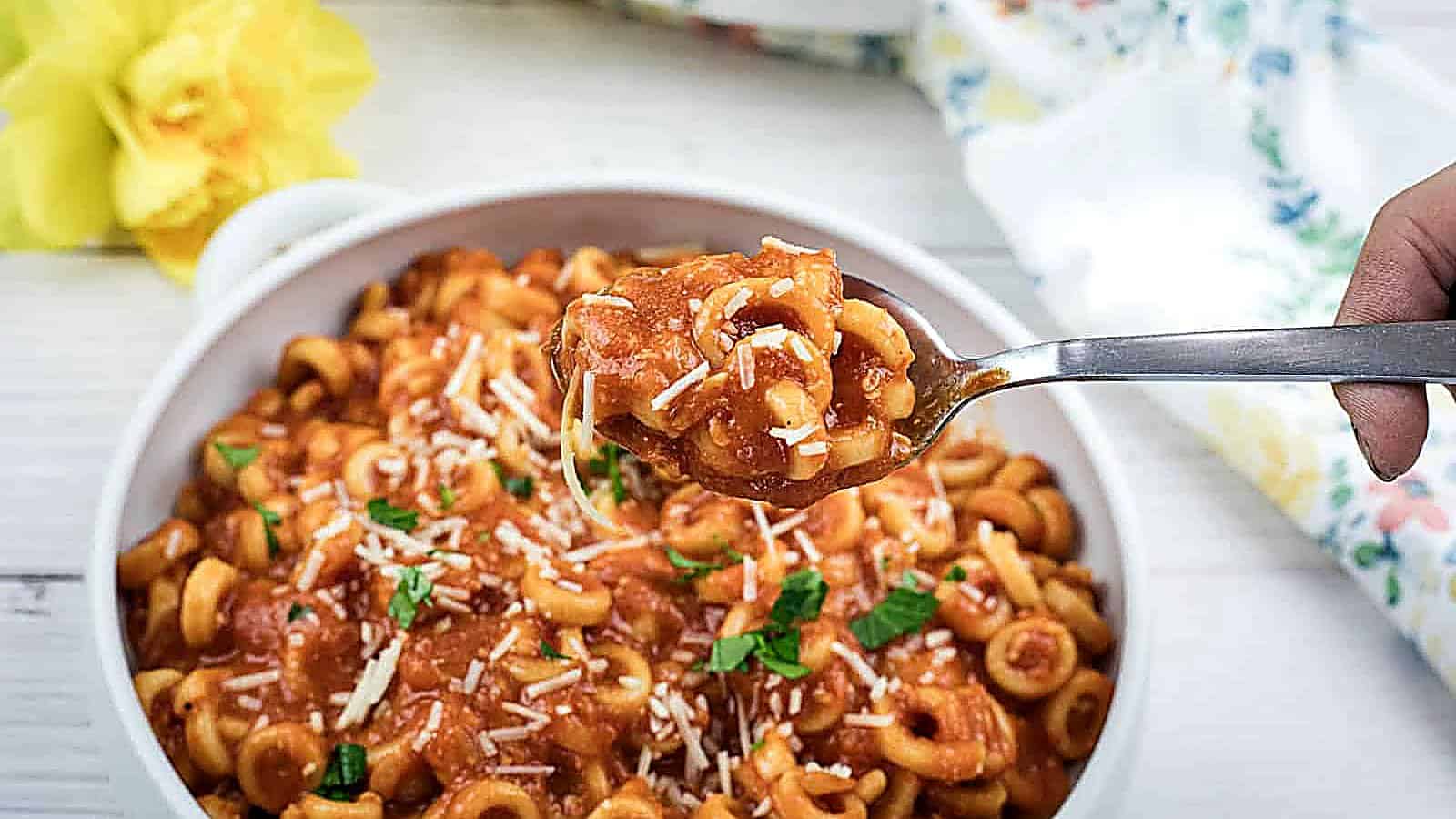 A bowl of pasta in tomato sauce, topped with grated chaeese and chopped herbs, with a spoon lifting a portion; a yellow flower and a floral napkin are nearby.