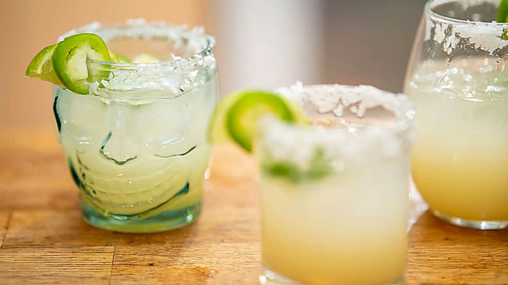 Three margaritas with salted rims and jalapeño slices, each showcasing unique margarita flavors, sit on a wooden surface. One glass is shaped like a skull.