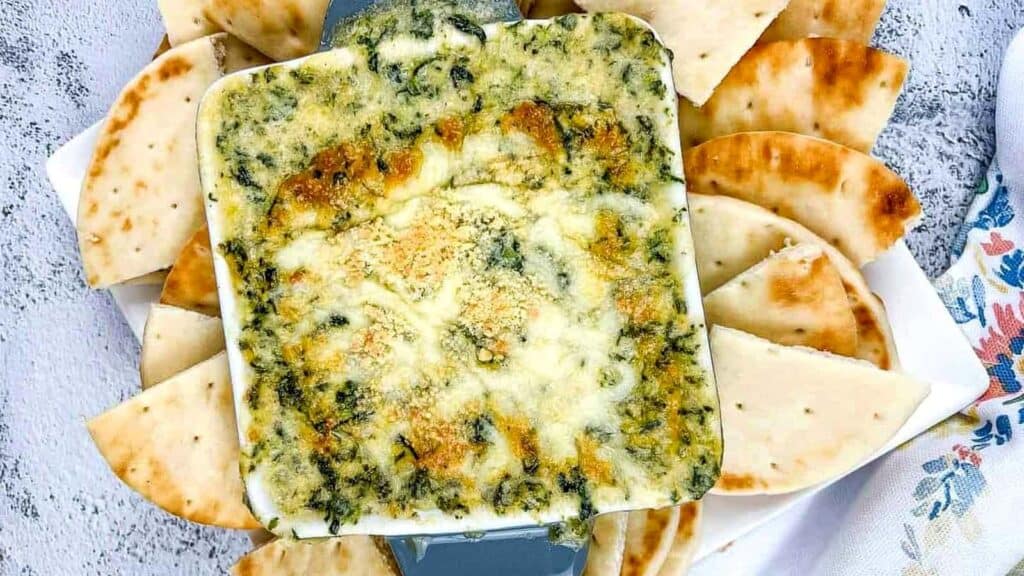 Baked spinach and cheese dip in a square dish, surrounded by slices of pita bread on a plate.