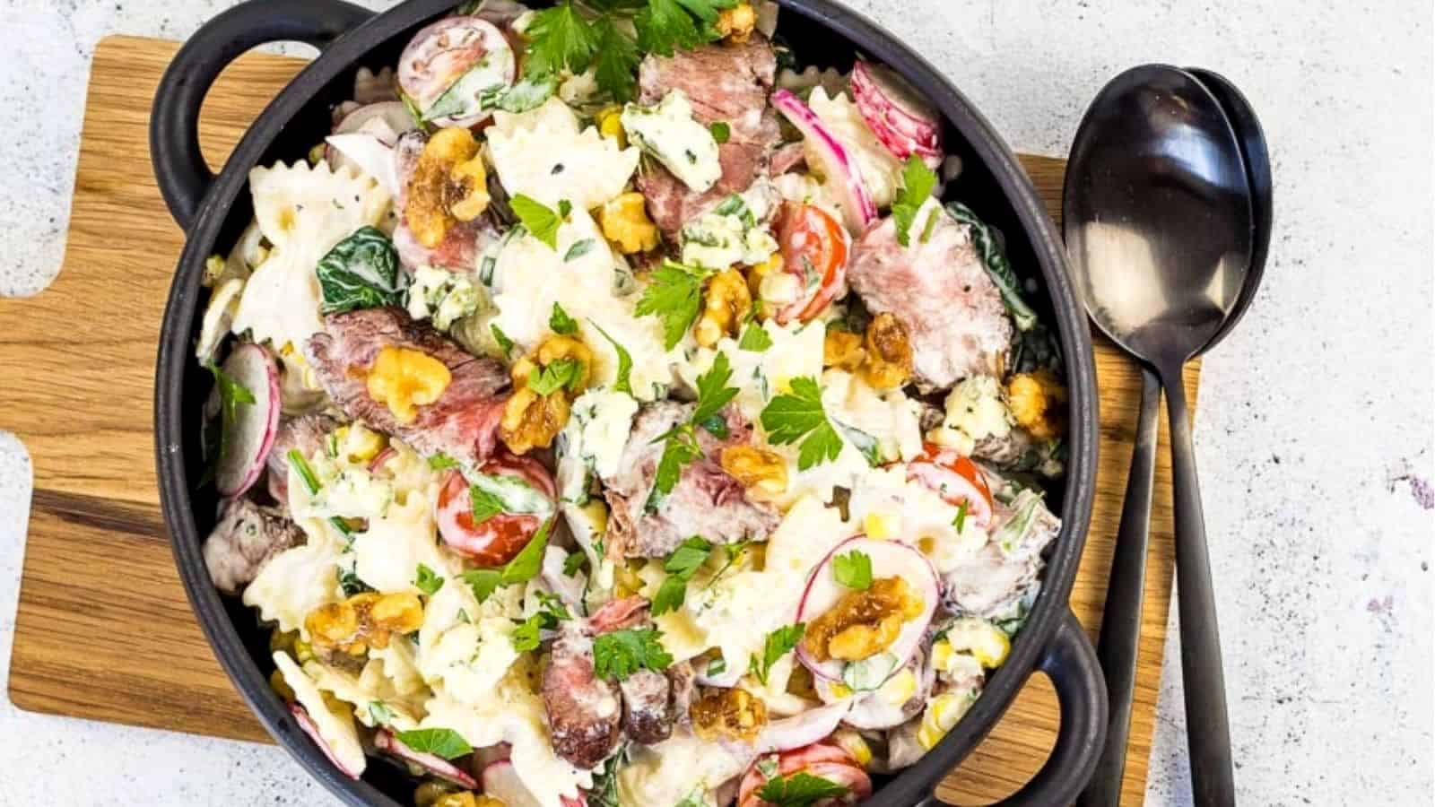 A black bowl filled with a pasta salad containing farfalle, steak slices, radishes, cherry tomatoes, lettuce, nuts, and crumbled blue cheese, garnished with parsley, on a wooden board next to a spoon.
