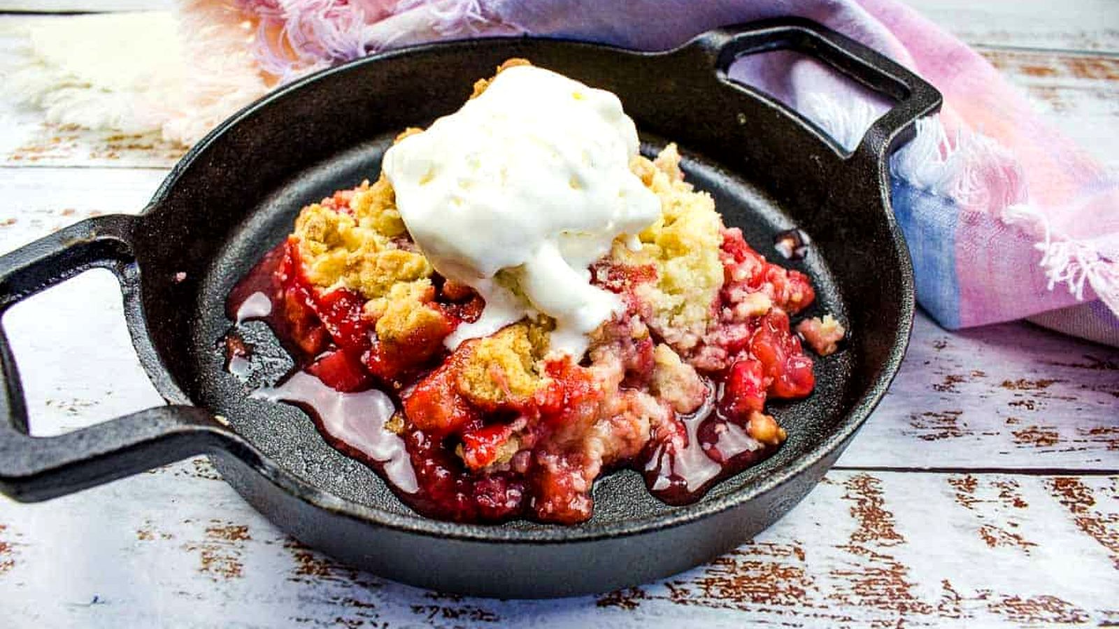 A cast iron skillet with strawberry rhubarb cobbler topped with a scoop of vanilla ice cream on a wooden surface with a cloth napkin beside it.