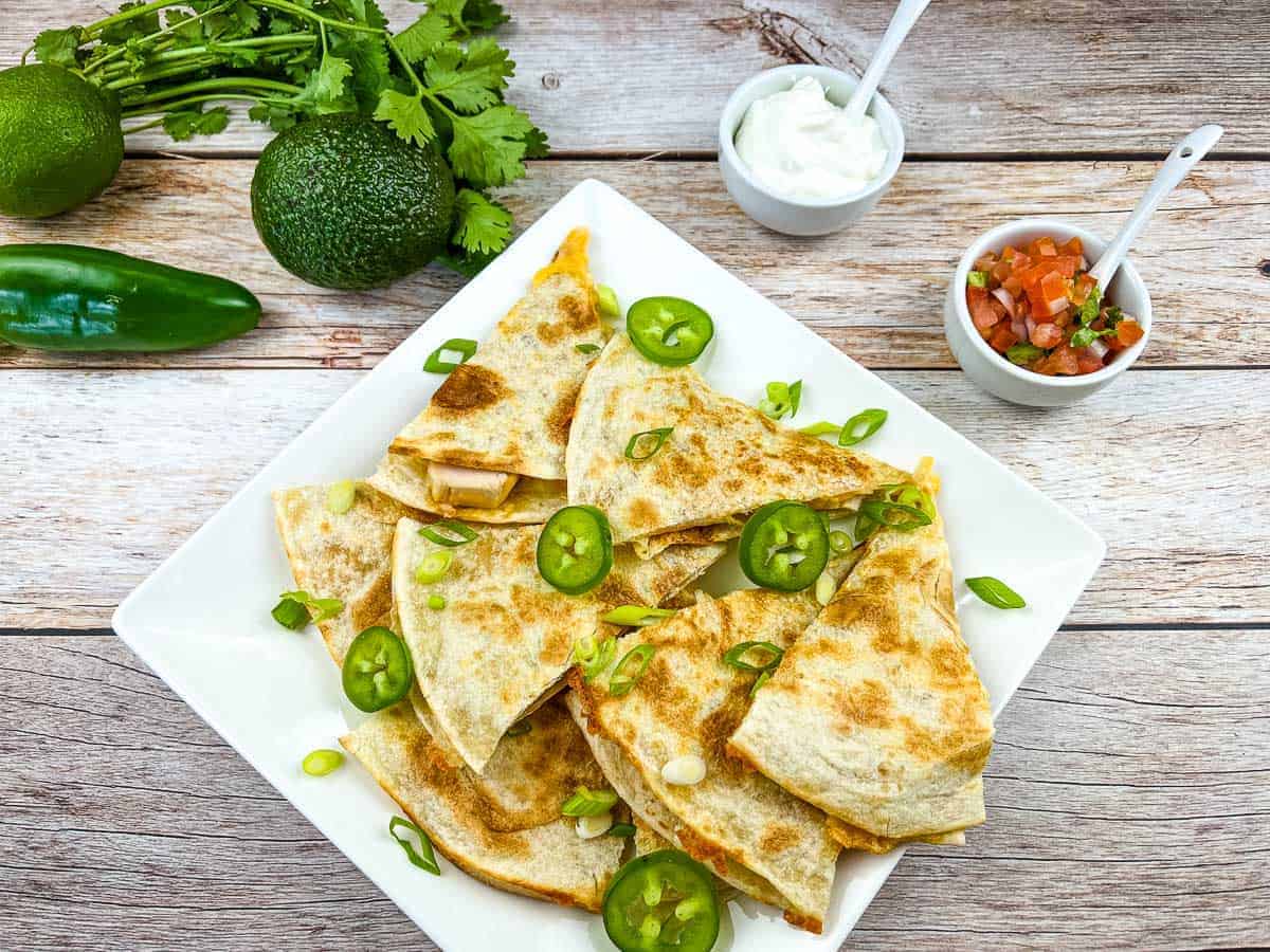 A plate of quesadillas garnished with sliced jalapeños and green onions, with sour cream, salsa, avocado, jalapeño, and cilantro on a wooden surface.