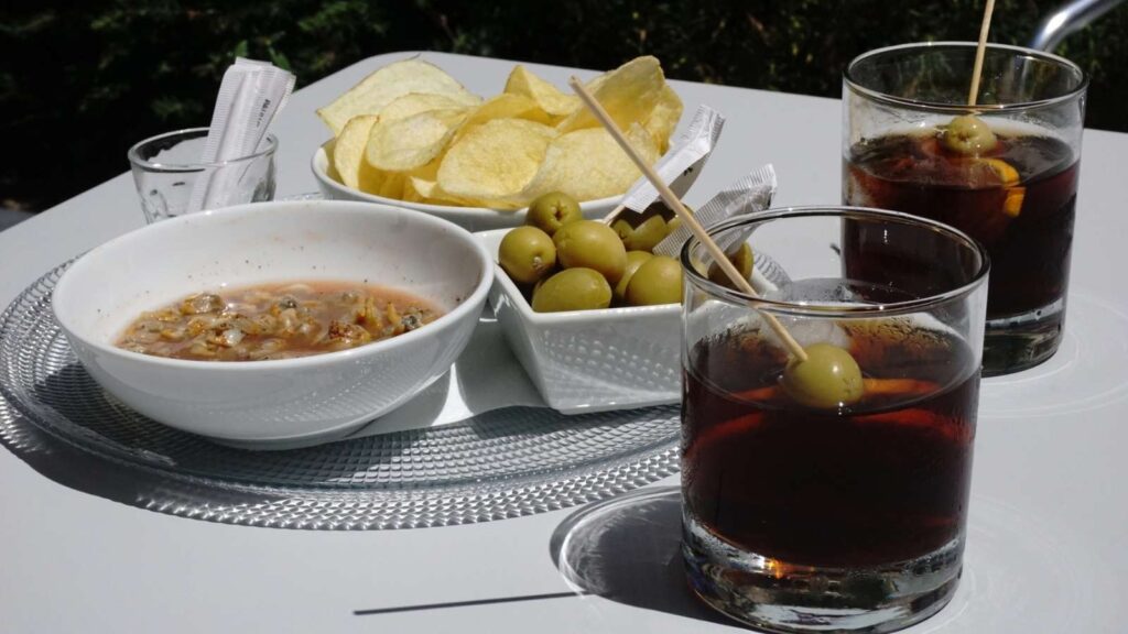 A table with two glasses of dark vermouth garnished with olives, a bowl of chips, a bowl of green olives, and a bowl of soup or stew, set outdoors.