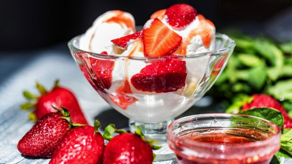 A glass bowl of vanilla ice cream sundae topped with sliced strawberries, surrounded by fresh strawberries and a small dish of strawberry syrup.