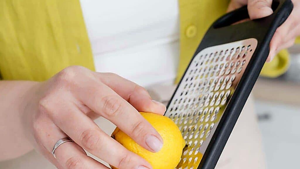 A person grates a lemon using a metal grater, holding the lemon with one hand and the grater with the other.