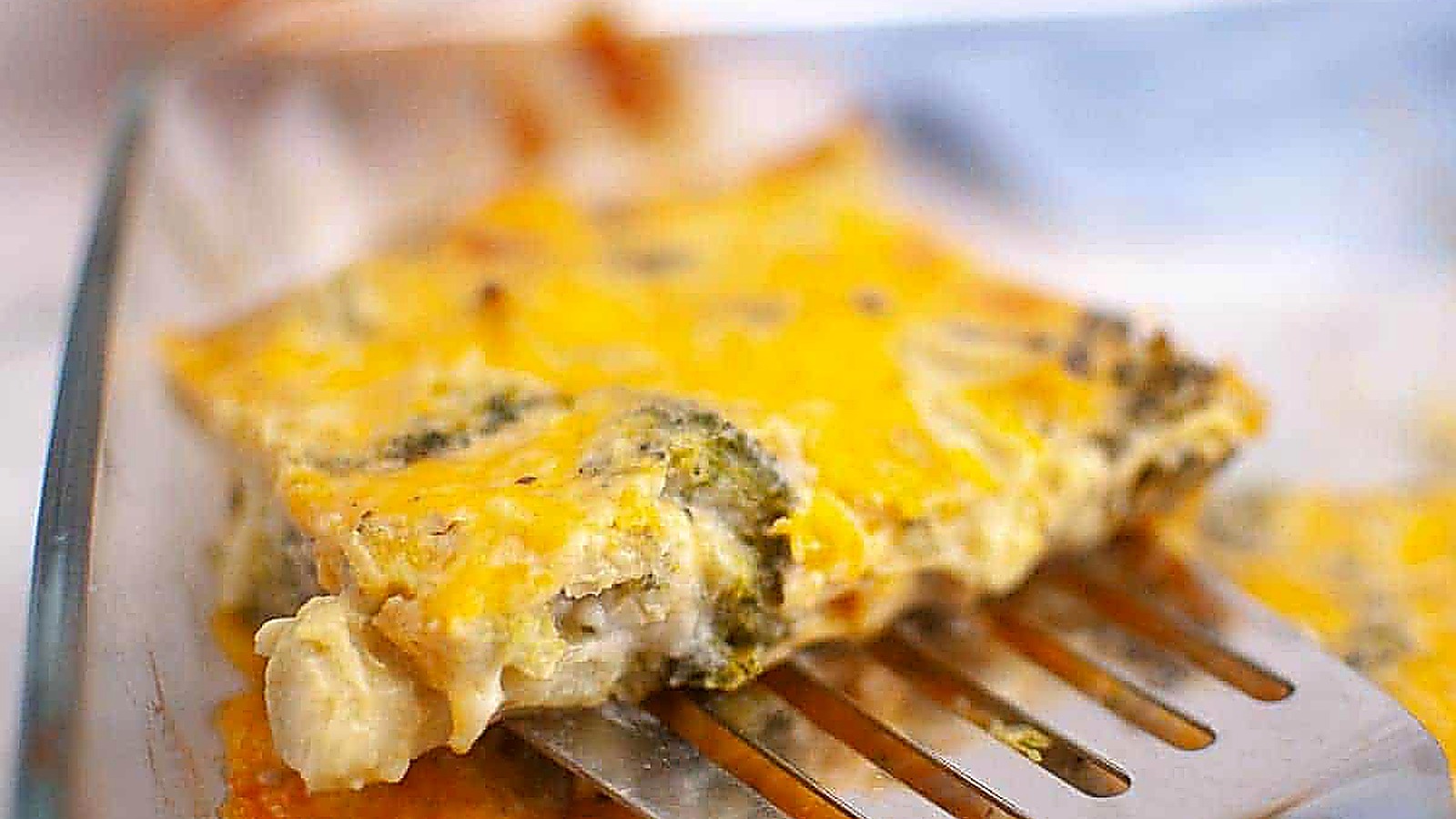 A metal spatula lifts a slice of 3-Ingredient Broccoli Cheese Casserole from a glass baking dish.
