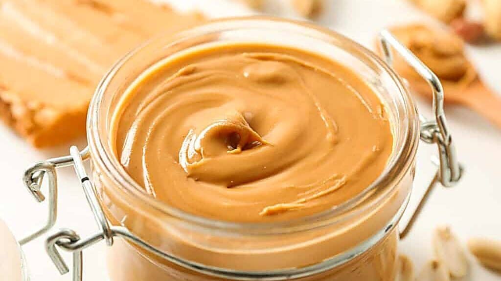 Open glass jar filled with creamy peanut butter, with a swirl pattern on the surface. Blurred peanuts and a slice of bread appear in the background.