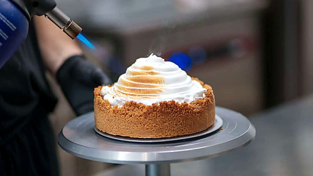 A person uses a kitchen torch to brown the meringue topping on a round pie with a crumb crust, placed on a rotating cake stand.