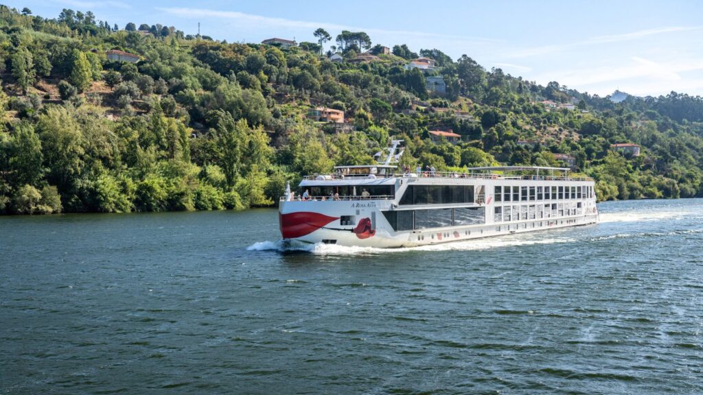 A white river cruise ship travels on a wide river, with green, tree-covered hills and scattered houses in the background under a clear sky.