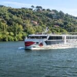 A white river cruise ship travels on a wide river, with green, tree-covered hills and scattered houses in the background under a clear sky.