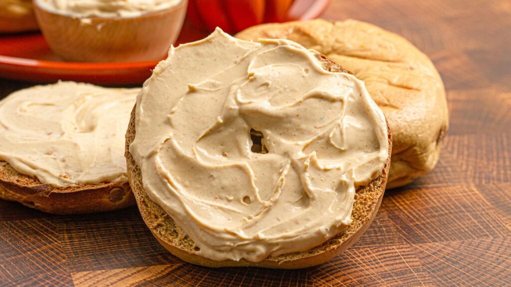 A toasted bagel half with a generous spread of light brown cream cheese sits on a wooden surface next to another bagel half and a plate in the background.
