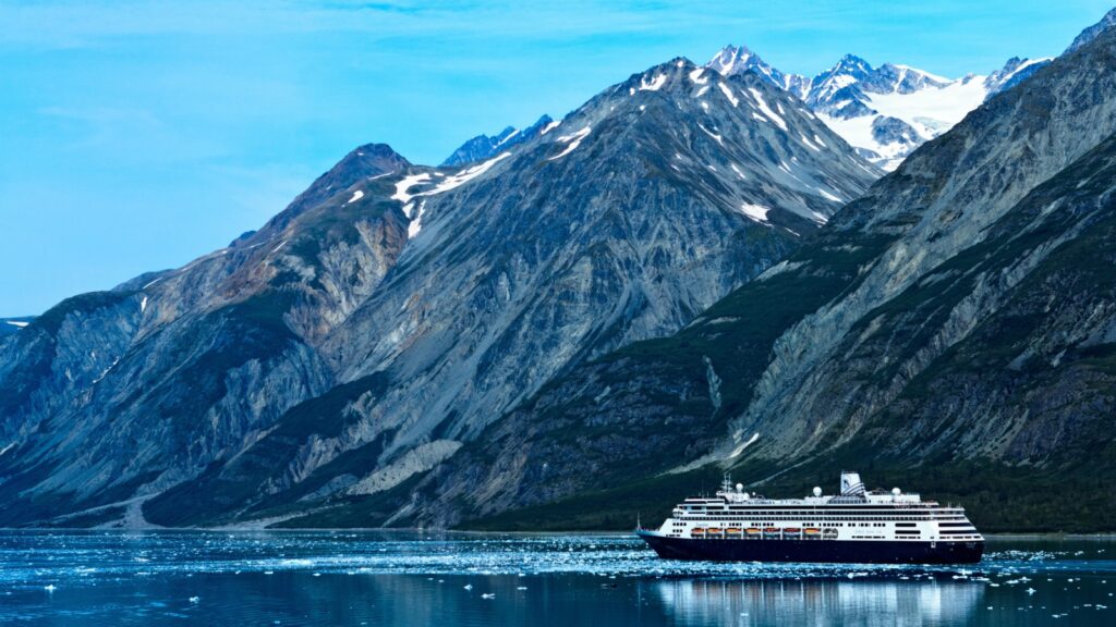A cruise ship sails through calm, icy waters with rugged, snow-capped mountains rising in the background under a clear blue sky.