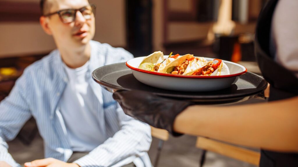 A server delivers a plate of tacos on a tray to a seated person at an outdoor restaurant, where regional chains rarely cross state lines, giving each location its own local flavor.