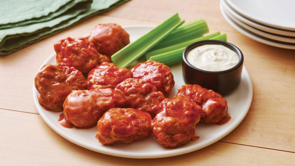 Plate of buffalo chicken wings with celery sticks and a cup of ranch dipping sauce, set on a wooden table next to a stack of white plates and a green napkin.