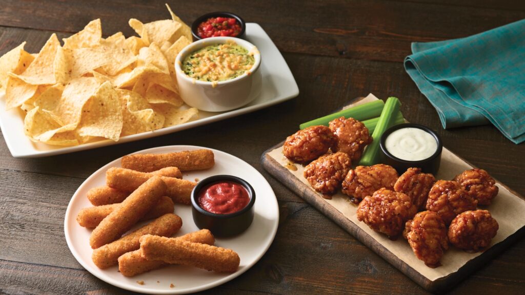 Plate of tortilla chips with dip, mozzarella sticks with marinara sauce, and boneless chicken wings with celery and ranch dressing on a wooden table.