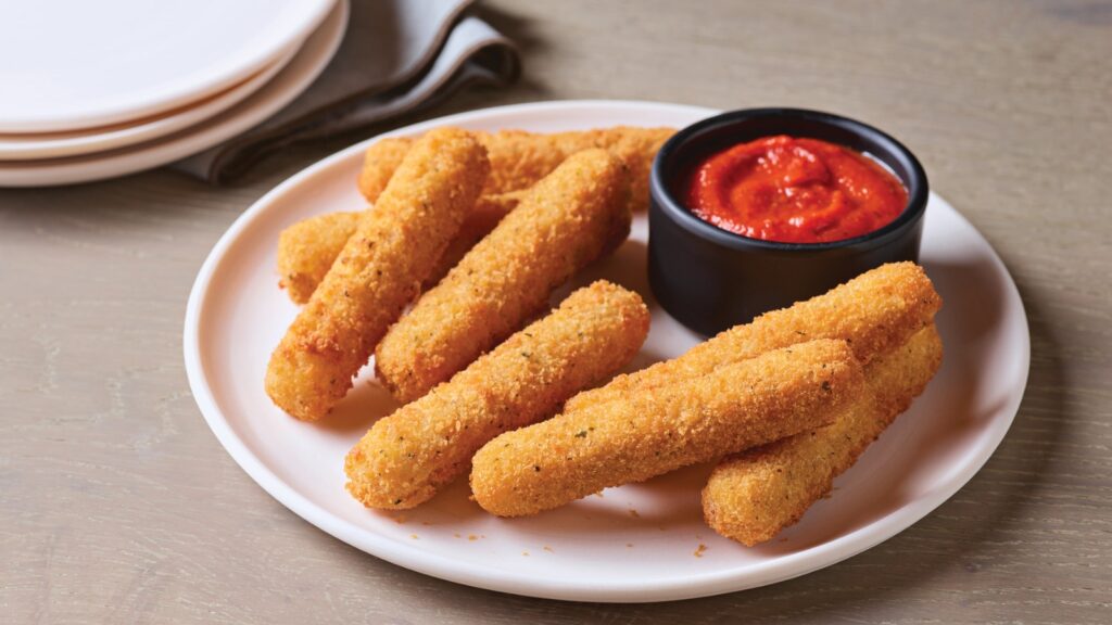 A white plate with breaded mozzarella sticks and a small black cup of marinara sauce, set on a light wooden table with stacked plates in the background.