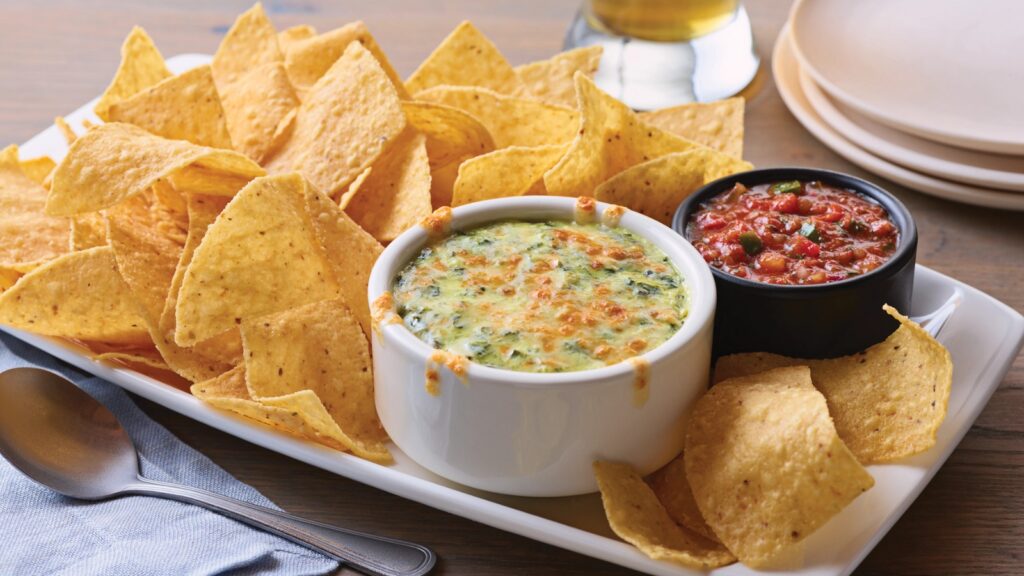 Plate of tortilla chips served with bowls of spinach cheese dip and tomato salsa, placed on a wooden table with a spoon and stacked plates nearby.