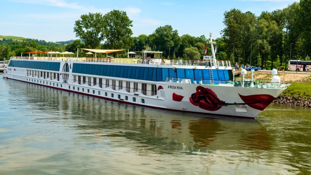 A long white river cruise ship with blue-tinted windows and a red lips graphic is sailing on a calm river near a green riverside with trees.