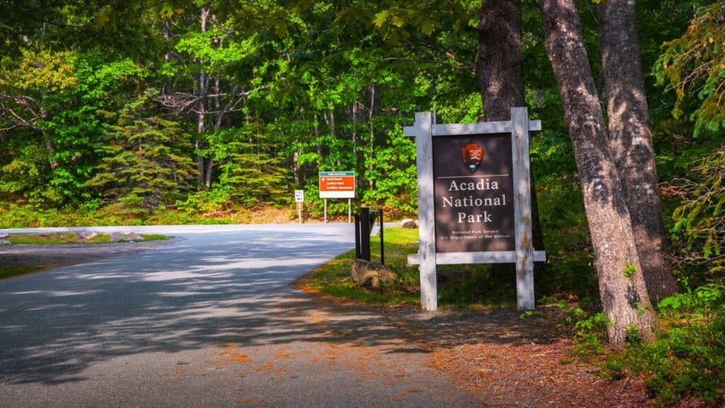 A large wooden sign reading “Acadia National Park” stands by a paved road surrounded by tall trees and dense green foliage on a sunny day—a picturesque reminder that, like many US national parks that require reservation, planning ahead can help you fully enjoy your visit.