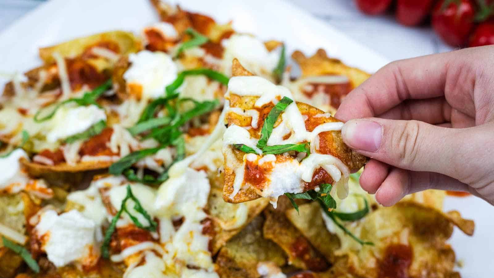 A hand holds a lasagna nachos topped with melted cheese, tomato sauce, ricotta, and basil over a plate of similar nachos.