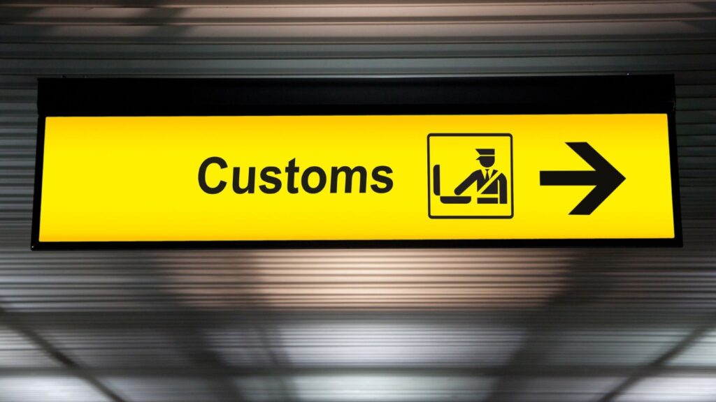 A yellow airport sign with the word &ldquo;Customs,&rdquo; an icon of an officer checking luggage, and a right arrow&mdash;reminding travelers to declare any items, including foods US customs will take, before proceeding.