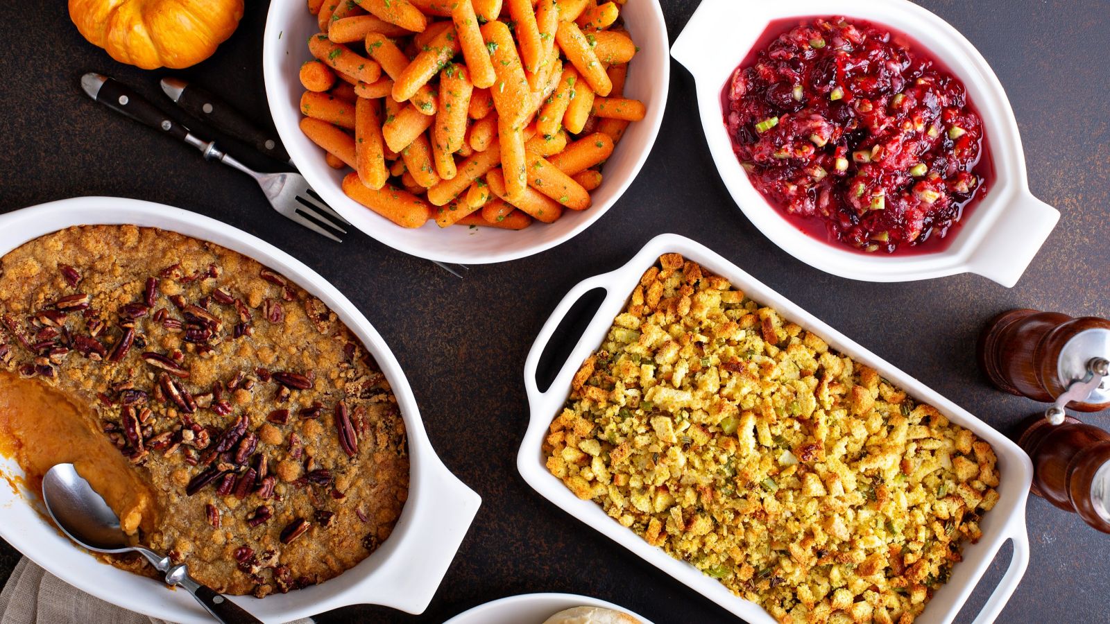A table with bowls of glazed carrots, cranberry sauce, pecan-topped casserole, and baked stuffing; utensils and condiments are also visible.