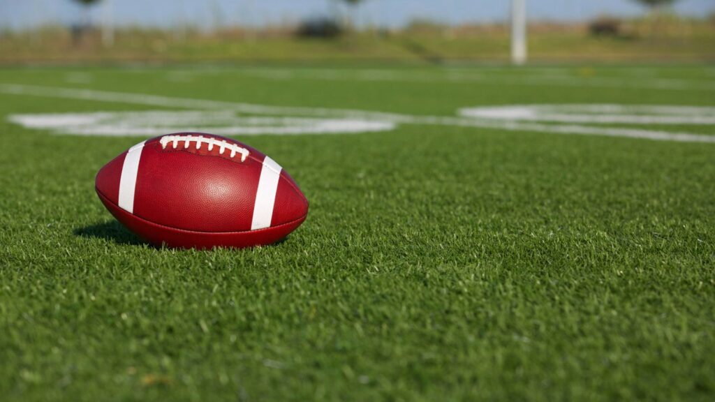 A football rests on the grass of an outdoor field with yard lines visible in the background.