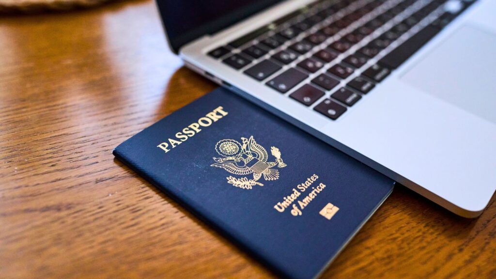 A United States passport rests partially underneath a silver laptop on a wooden table.