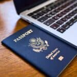 A United States passport rests partially underneath a silver laptop on a wooden table.