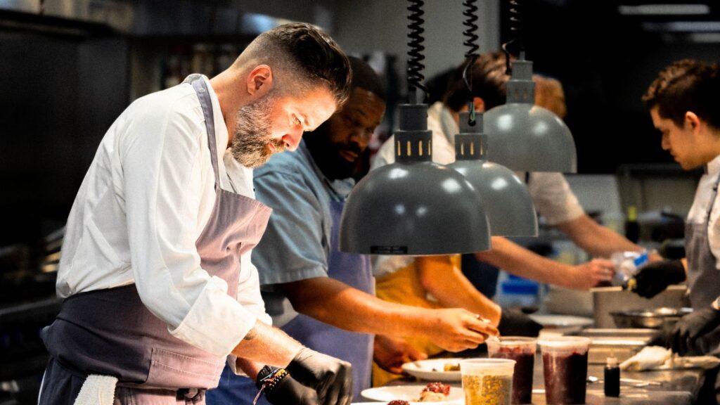 Four chefs prepare and plate food under pendant lights in a busy commercial kitchen, each focused on their individual tasks.