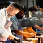 Four chefs prepare and plate food under pendant lights in a busy commercial kitchen, each focused on their individual tasks.