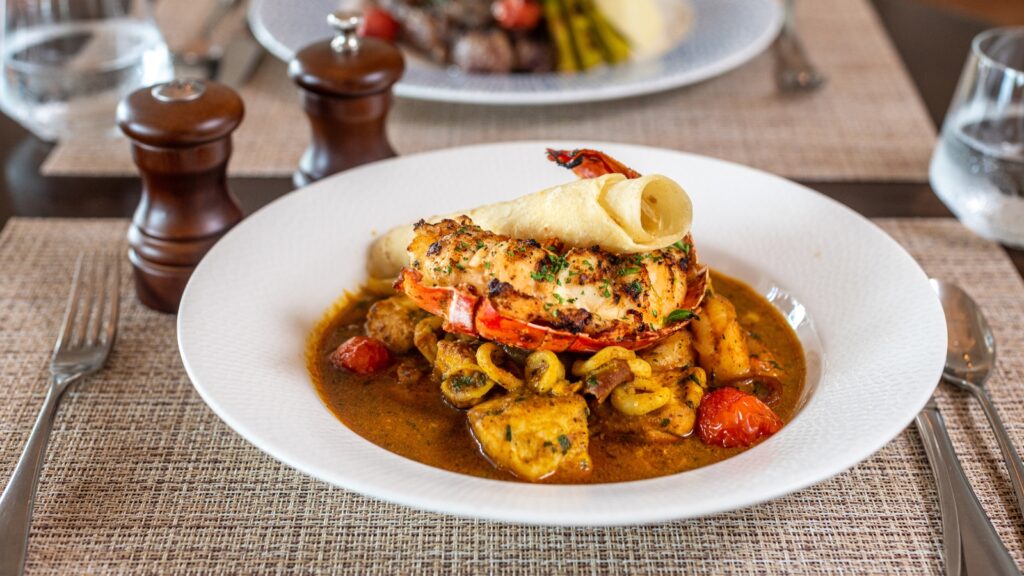 A white plate with lobster tail, rolled crepe, assorted seafood, cherry tomatoes, and sauce, placed on a woven placemat with cutlery, water glasses, and salt and pepper shakers.