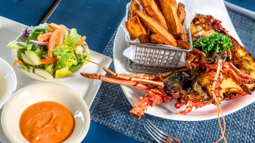 Grilled lobster with fries in a basket, mixed salad, and dipping sauces served on a blue placemat.
