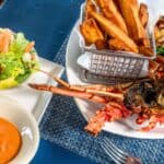Grilled lobster with fries in a basket, mixed salad, and dipping sauces served on a blue placemat.