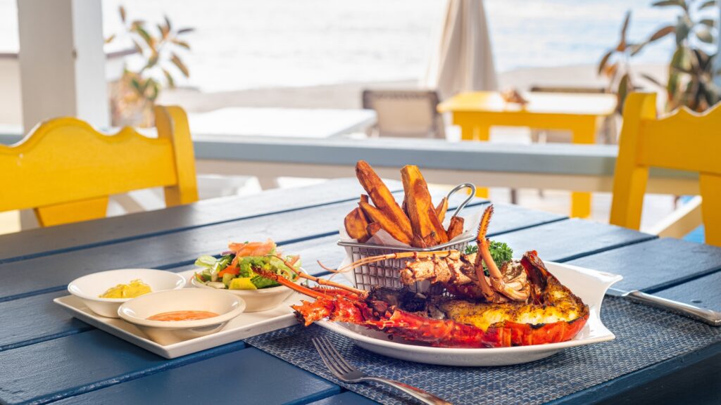 Lobster, fries, dipping sauces, and salad served on a blue table at an outdoor seaside restaurant with yellow chairs.
