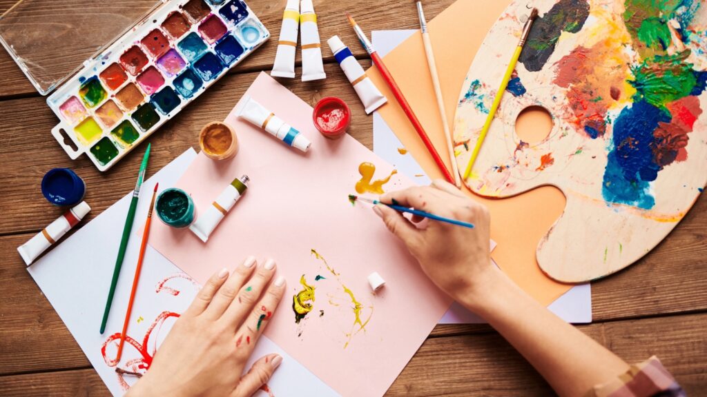 Person painting on paper with a paintbrush, surrounded by paints, brushes, palette, and art supplies on a wooden table.