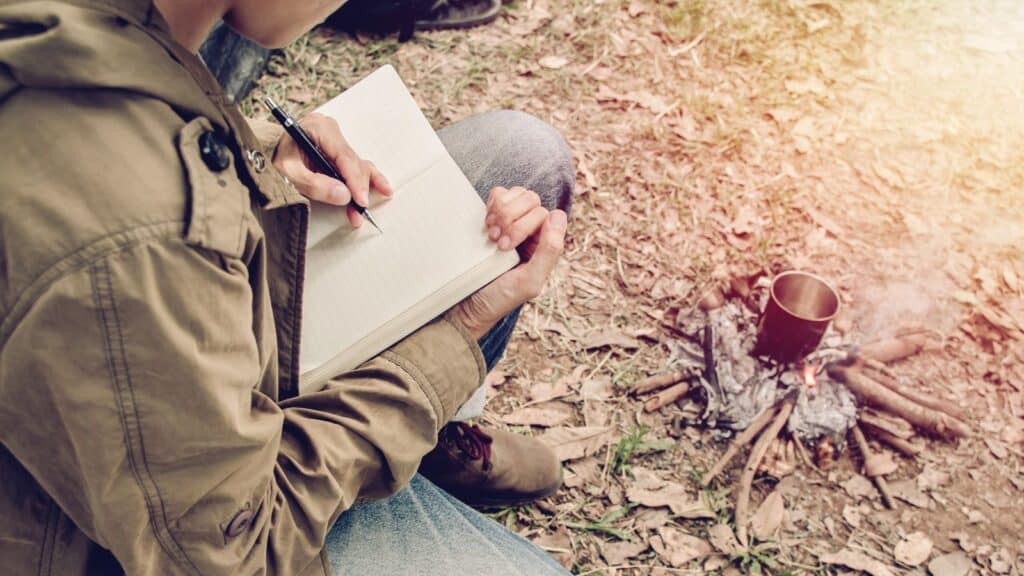 A person in a brown jacket writes in a notebook while sitting near a small campfire with a metal cup heating on it, outdoors on leaf-covered ground&mdash;capturing the summer camp traditions campers remember most fondly.