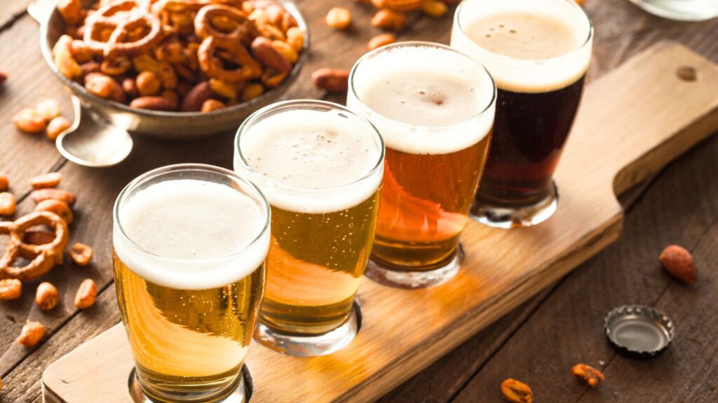 Four small glasses of assorted beer on a wooden flight board, with a bowl of mixed snacks and pretzels in the background on a wooden table.