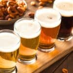 Four small glasses of assorted beer on a wooden flight board, with a bowl of mixed snacks and pretzels in the background on a wooden table.