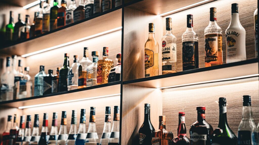 Shelves filled with various liquor bottles, including vodka, rum, whiskey, and liqueurs, organized in rows under warm lighting in a bar setting.