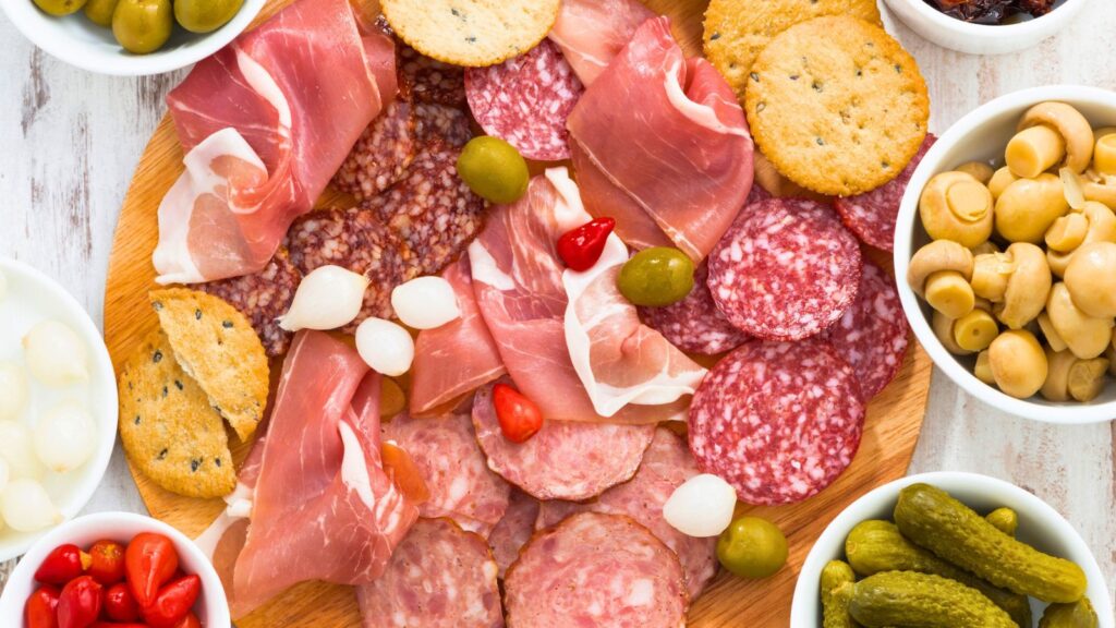 A charcuterie board with assorted cured meats, crackers, olives, onions, pickles, small peppers, mushrooms, and bread slices on a wooden surface offers the perfect setup for savory snacking, surrounded by small bowls.