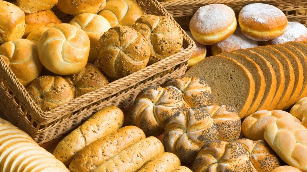 Assorted breads including rolls, baguettes, seeded buns, sliced rye, and powdered doughnuts&mdash;favorites among Americans&mdash;are displayed in baskets and on a table. These tasty offerings highlight the variety enjoyed here compared to the stricter EU rules governing bakery products.