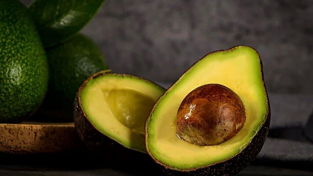 A halved avocado with the seed visible is in the foreground, while whole avocados and leaves are in the background on a dark surface.
