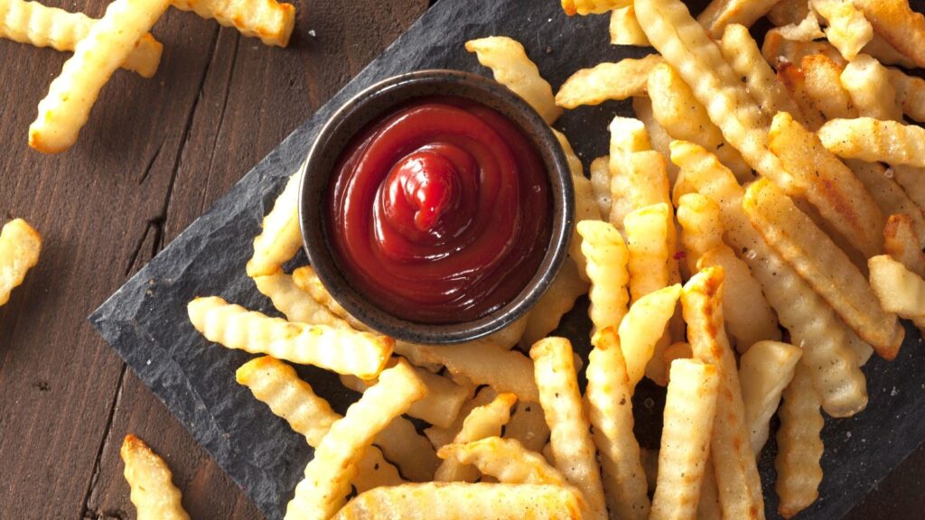 Crinkle-cut French fries arranged around a small bowl of ketchup on a dark slate serving tray, placed on a wooden surface.
